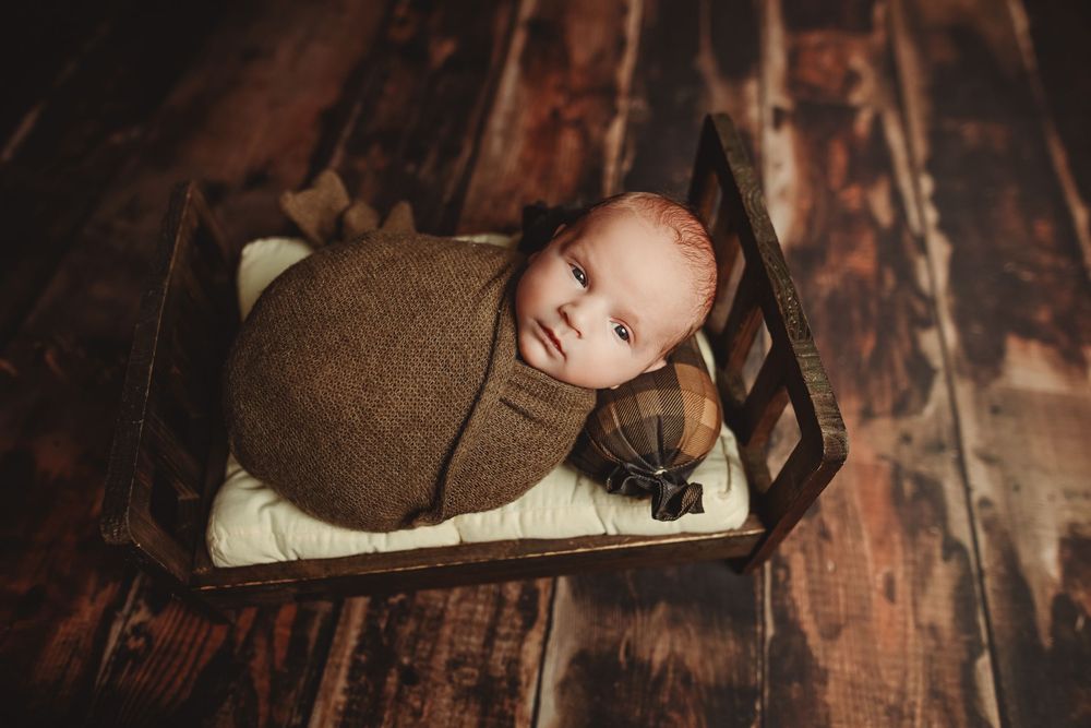 Newborn baby swaddled in brown wrap sleeping peacefully in a tiny wooden bed on rustic hardwood floors.
