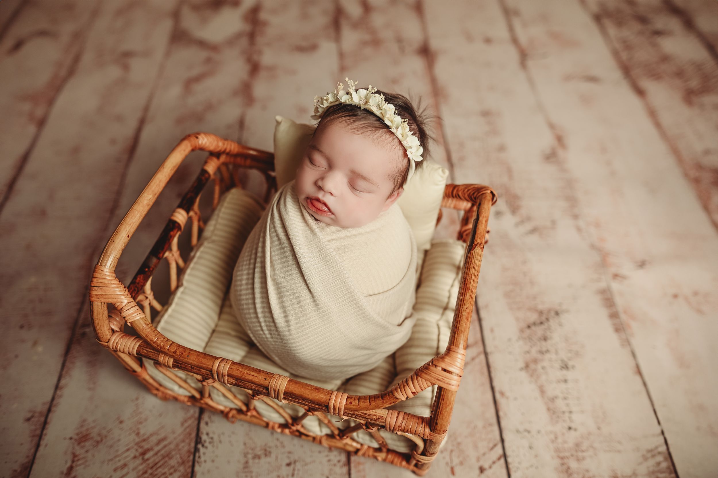 Newborn baby wrapped in cream swaddle with floral headband sleeping peacefully in a wicker basket on wood floor.