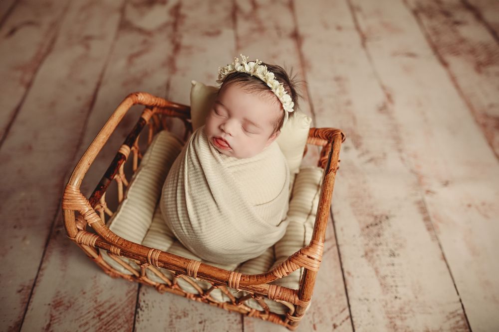 Newborn baby wrapped in cream swaddle with floral headband sleeping peacefully in a wicker basket on wood floor.