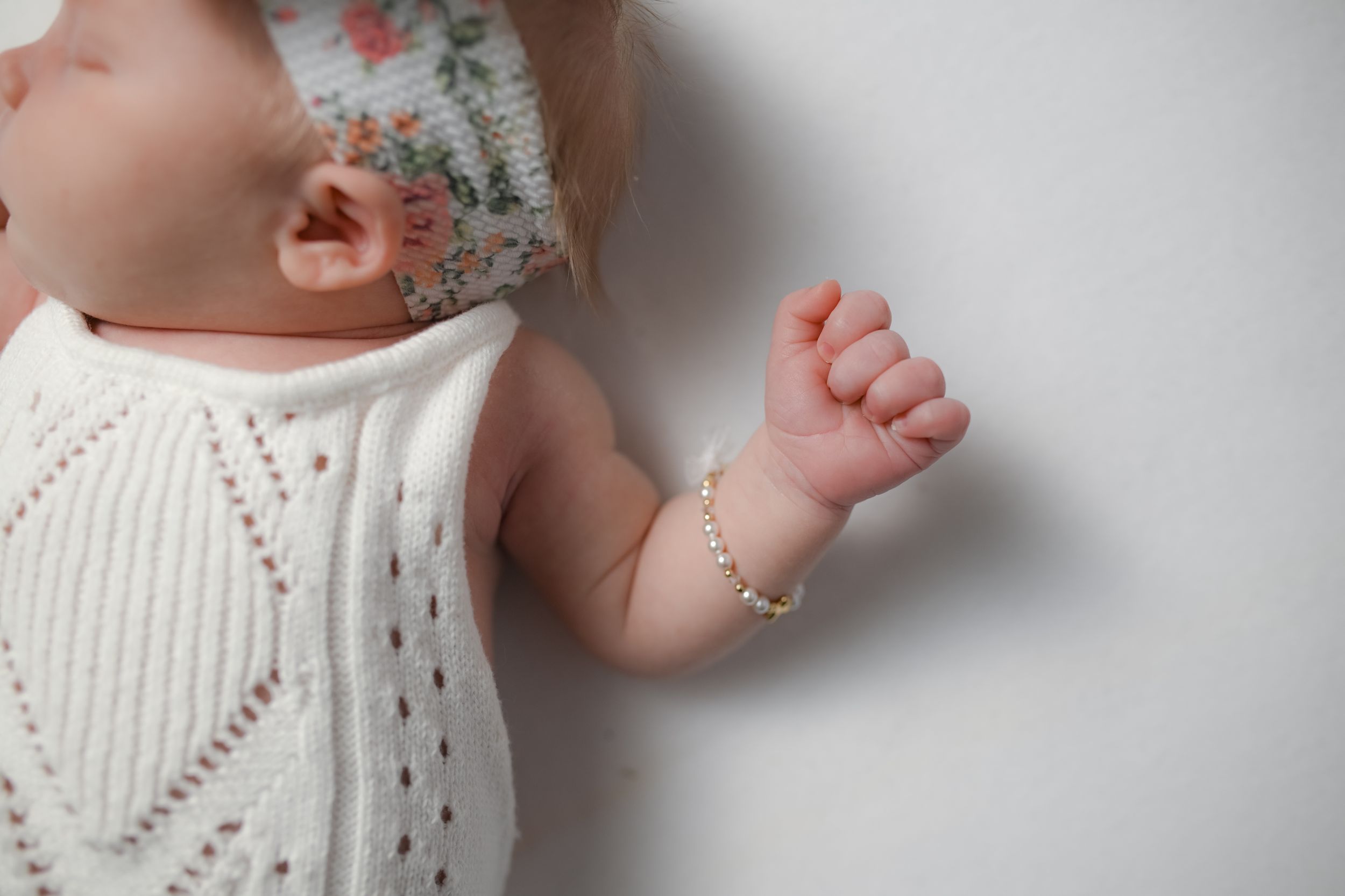 Newborn baby wearing a white knit romper and delicate pearl bracelet with a floral headband, lying on white background.