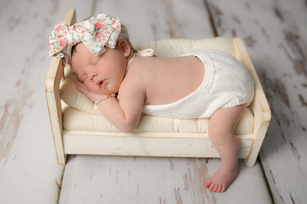 Newborn baby girl sleeping peacefully in a tiny wooden bed, wearing a white romper and floral bow headband.