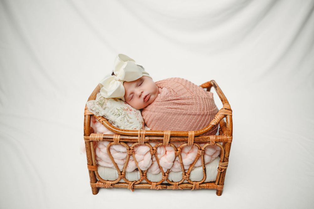 Newborn baby resting in coral wrap on wicker doll bed against white background.