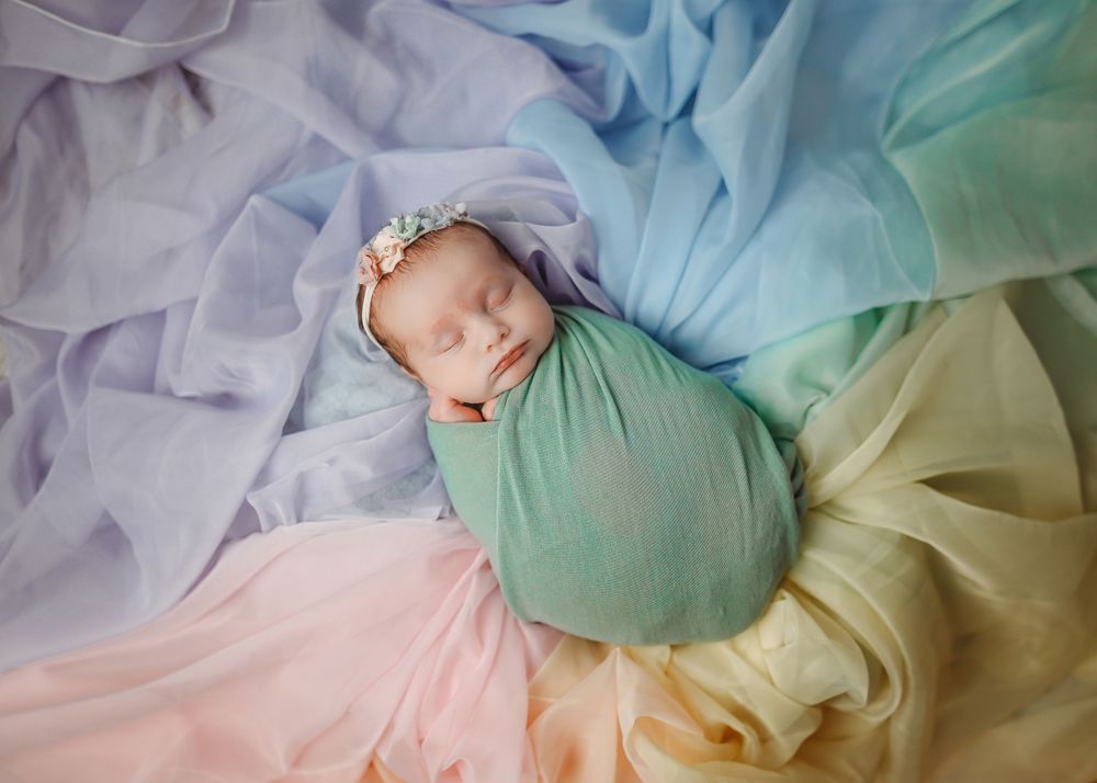 Sleeping newborn baby swaddled in green wrap on colorful pastel rainbow fabric backdrop.