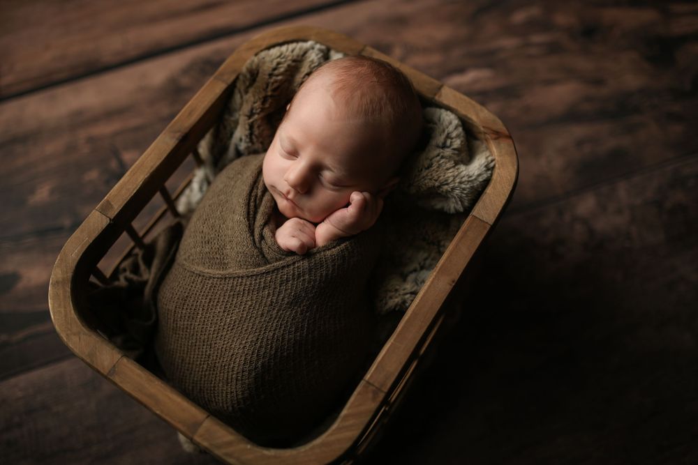 Newborn baby swaddled in olive green wrap, sleeping peacefully in a rustic wooden basket on dark wood floor.