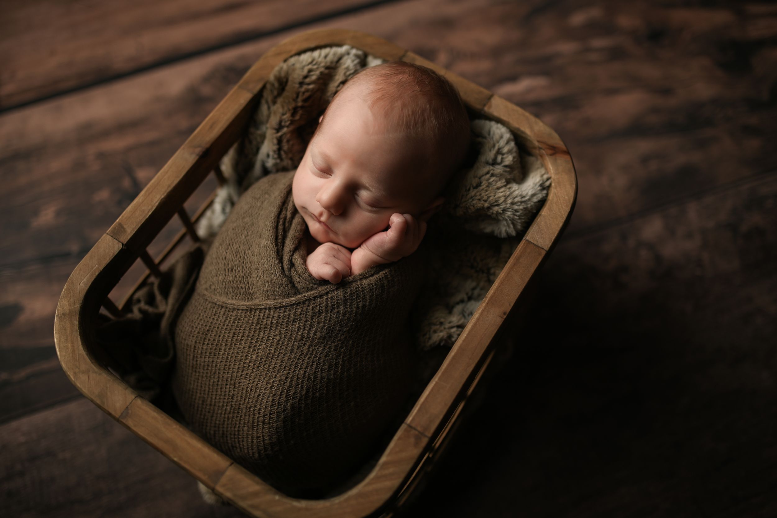 Newborn baby swaddled in olive green wrap, sleeping peacefully in a rustic wooden basket on dark wood floor.