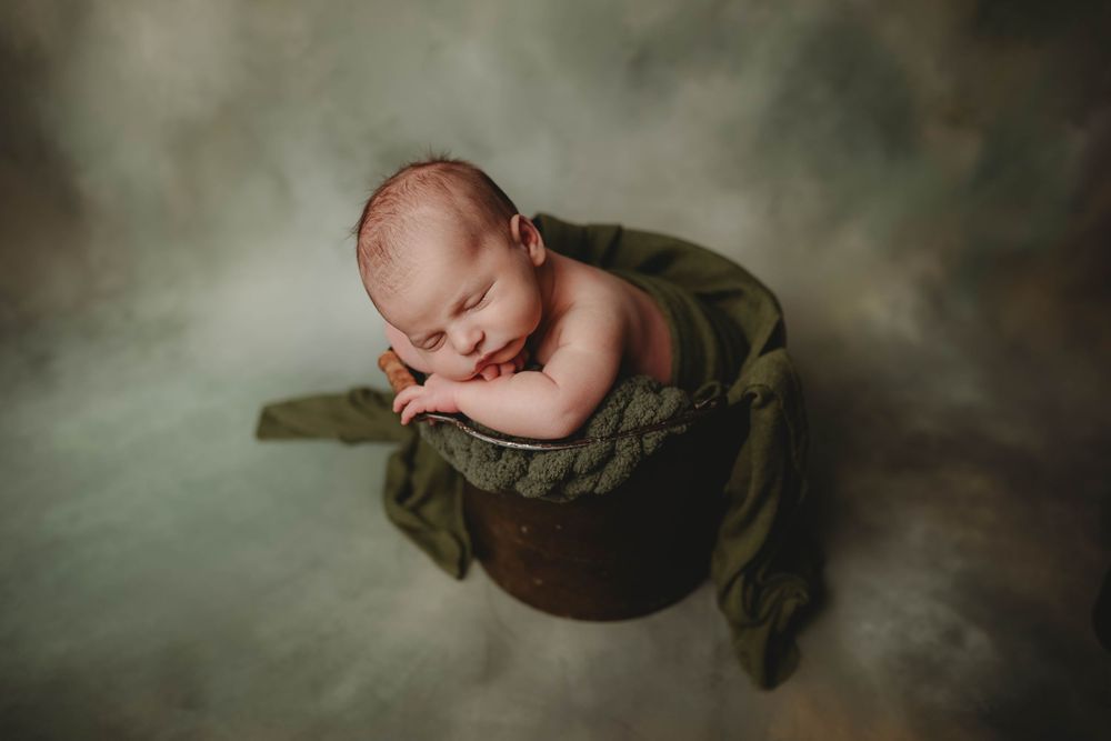 Sleeping newborn baby curled in a rustic wooden bucket with green fabric on a soft gray background.