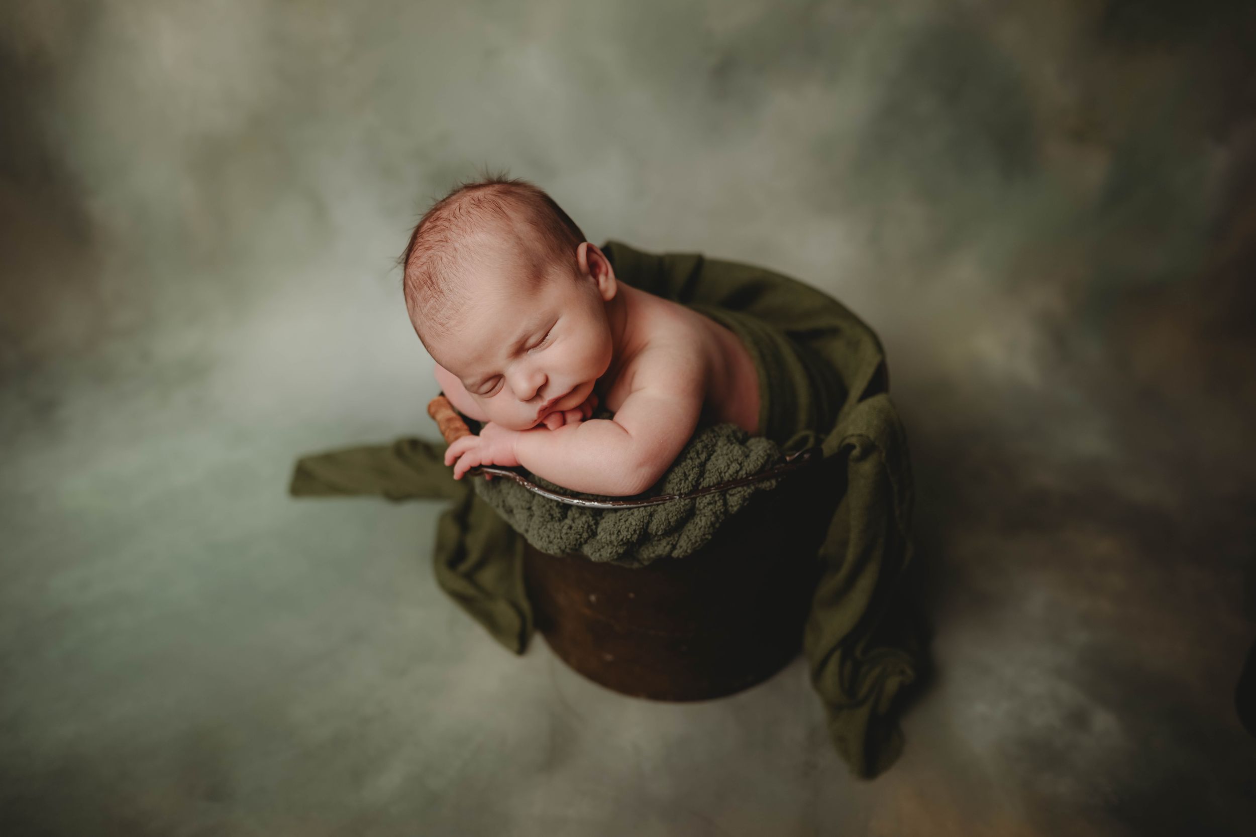Sleeping newborn baby curled in a rustic wooden bucket with green fabric on a soft gray background.