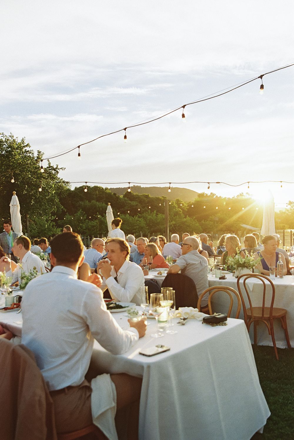 Outdoor reception event with string lights overhead and guests dining at white-clothed tables during golden hour sunset.