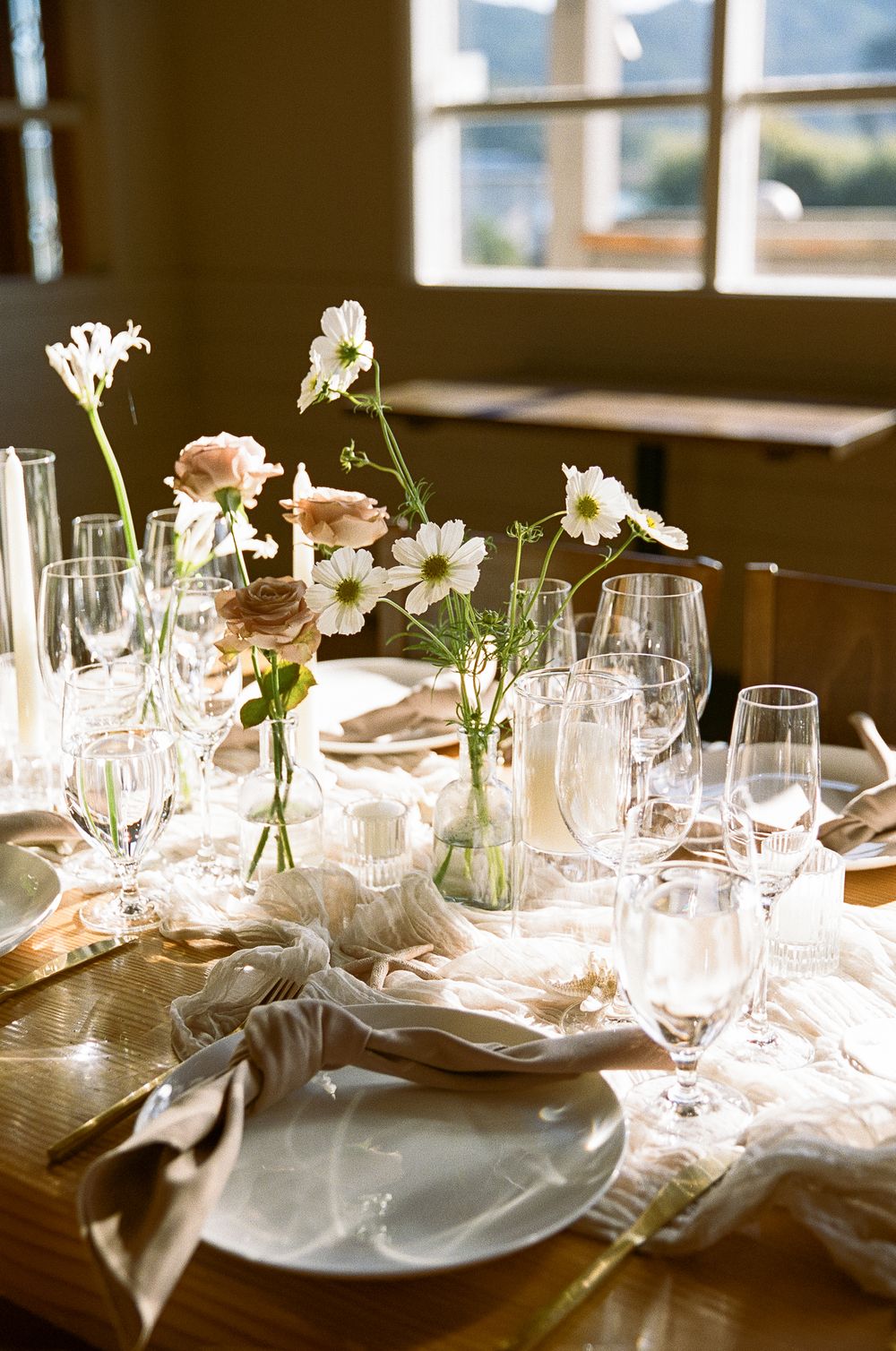 An elegant table setting with wine glasses and delicate white flowers in vases near sunlit windows.