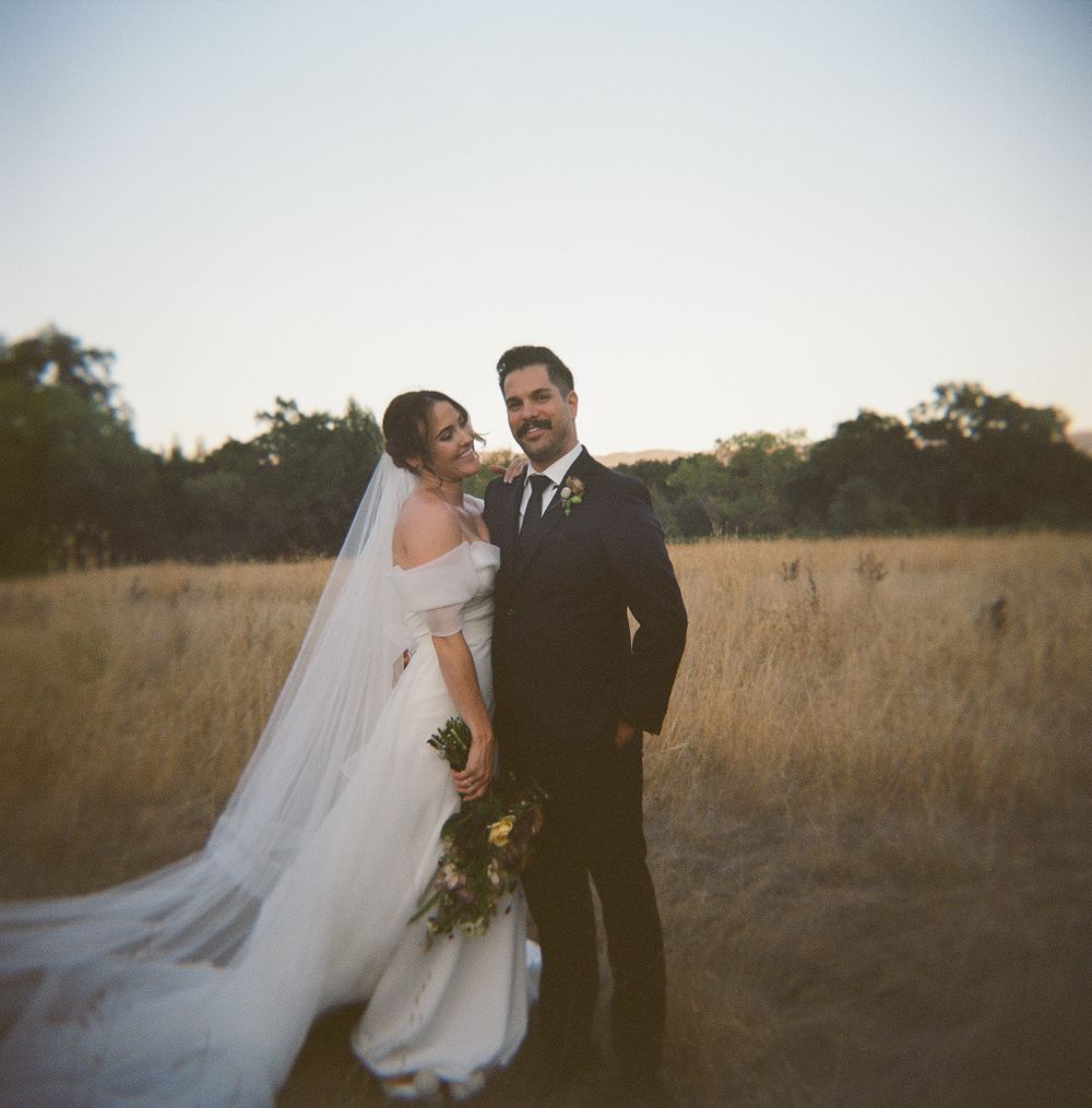 A couple in wedding attire stand together in a golden field at sunset, with a long flowing white veil trailing behind.