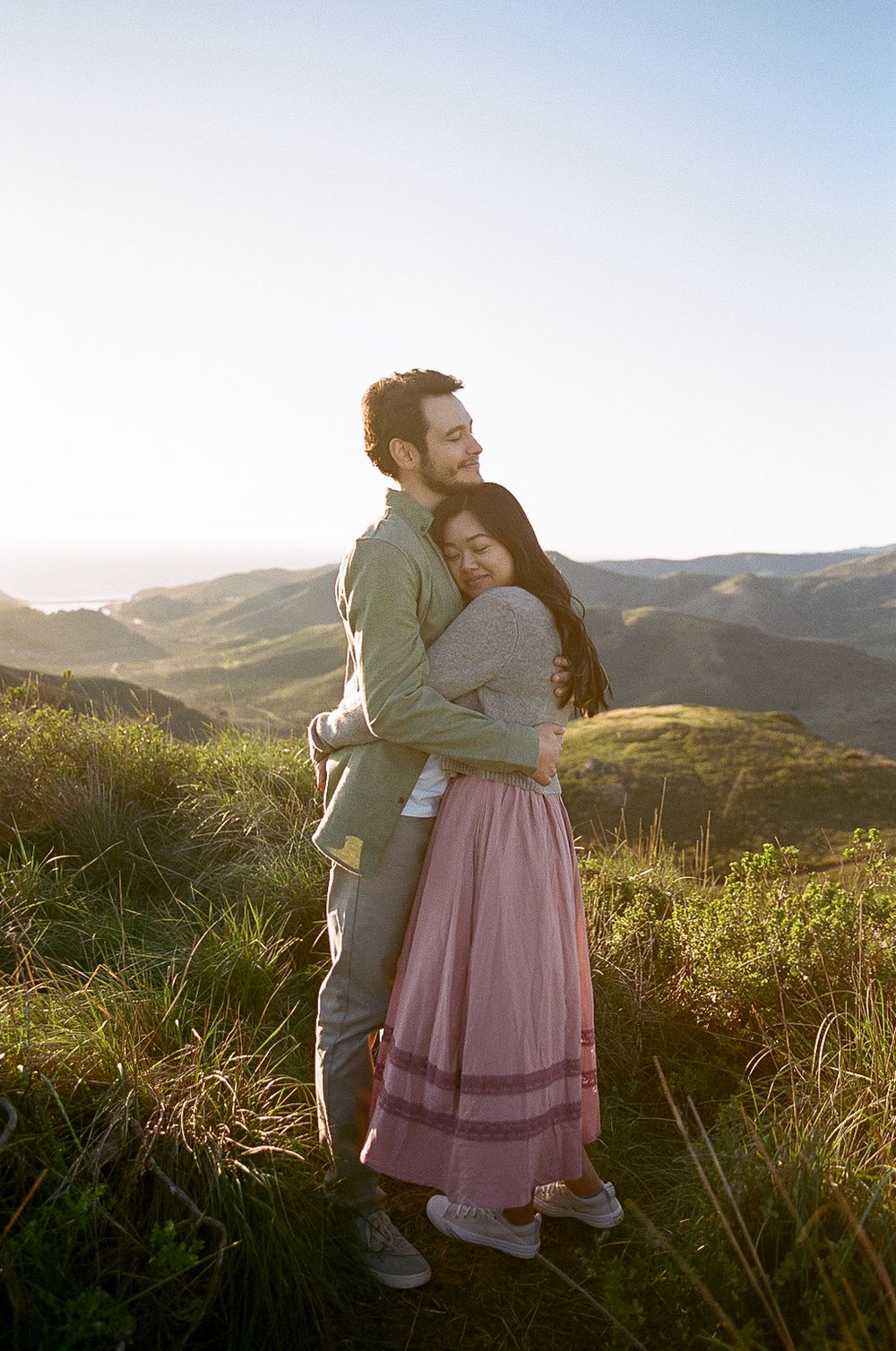 A couple embraces on a hilltop during golden hour as the sun sets over rolling mountains in the background.