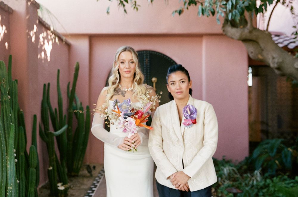 A couple in wedding attire poses together against a pink wall with tall cacti and greenery in the background.
