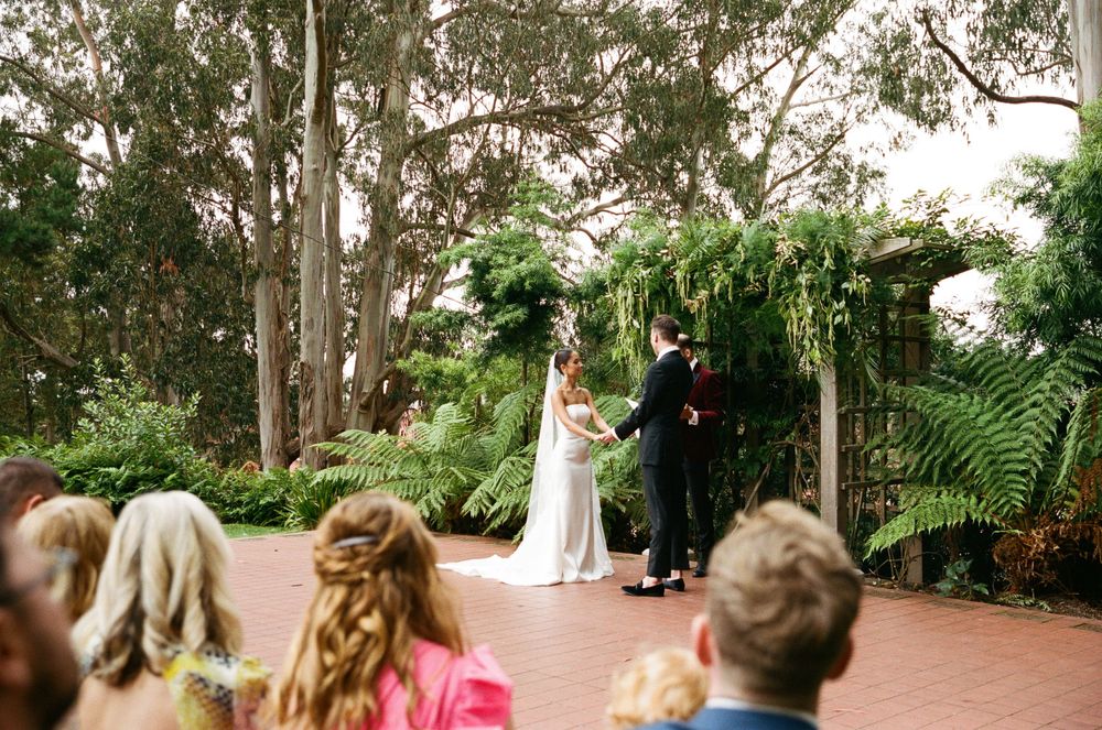 A wedding ceremony takes place outdoors on a brick patio surrounded by lush tropical gardens and tall eucalyptus trees.