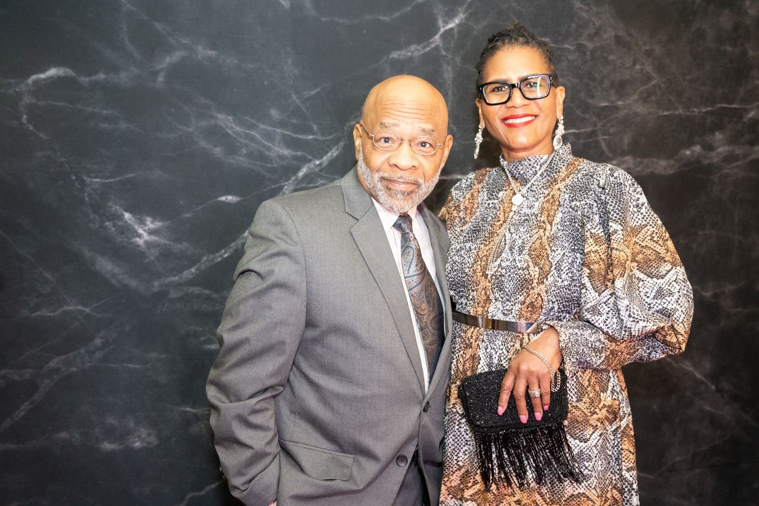 A couple dressed in formal attire pose together in front of a dark marbled backdrop.