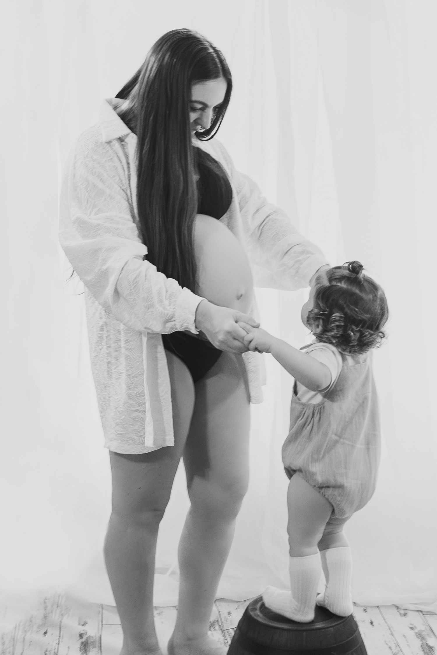 A tender black and white maternity moment shows a pregnant woman in white holding hands with a small toddler.