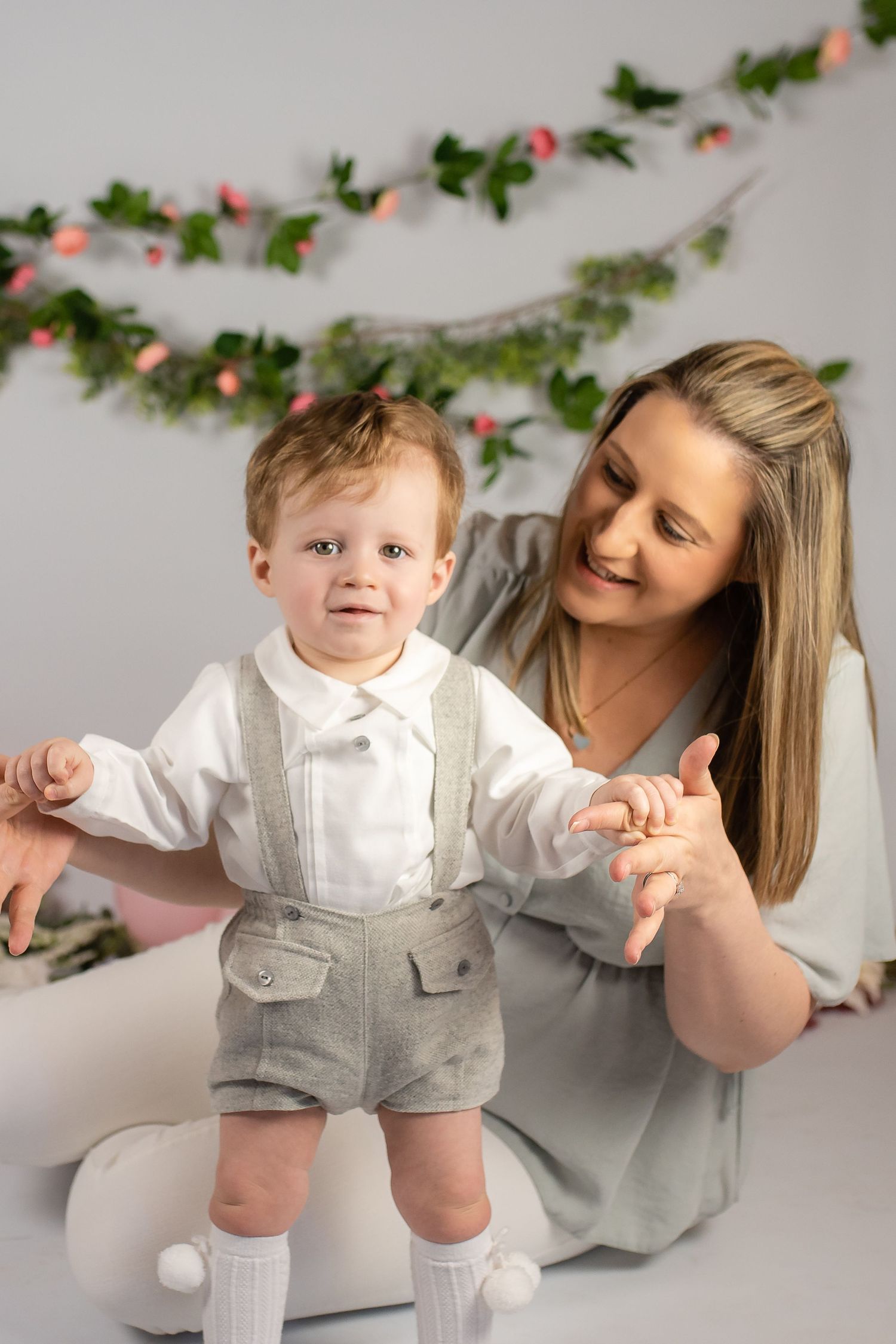 Sweet holiday photo scene with a small boy in suspender shorts and bow tie against a floral garland backdrop.