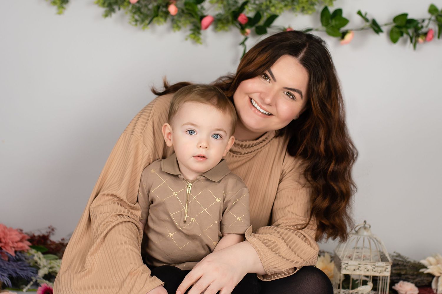 A parent and child sit together in matching tan outfits against a white wall decorated with holiday garland and florals.