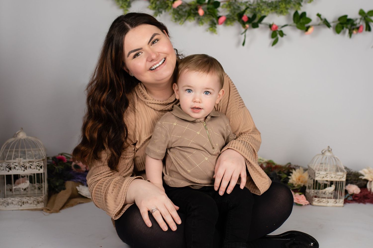 A parent and young child wearing matching tan sweaters sit together on the floor with holiday decorations in the background.