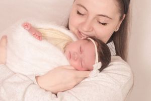 A tender moment shows a sleeping newborn baby in a soft white outfit being cradled lovingly against someone's chest.