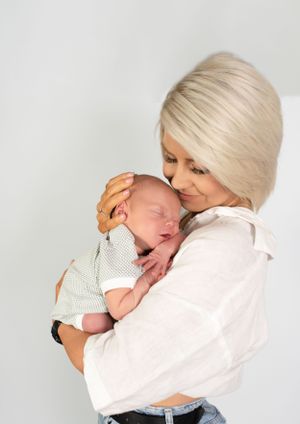 A tender portrait of a woman in white holding and kissing a sleeping newborn baby.