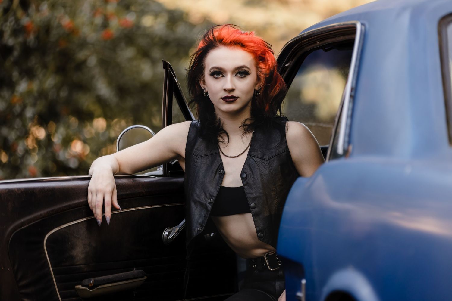 A figure with vibrant red hair sits in a vintage car wearing a black vest and dark makeup in an autumn setting.