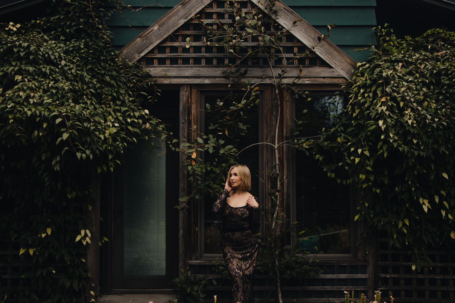 Moody portrait of a woman in dark attire standing in front of an ivy-covered garden entrance.