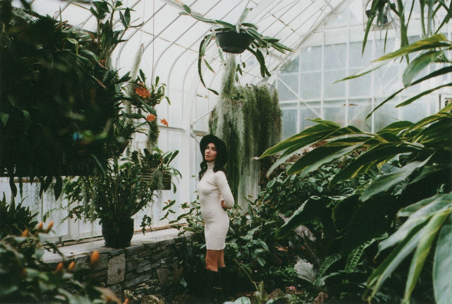 Woman in white dress standing among tropical plants inside a large greenhouse.