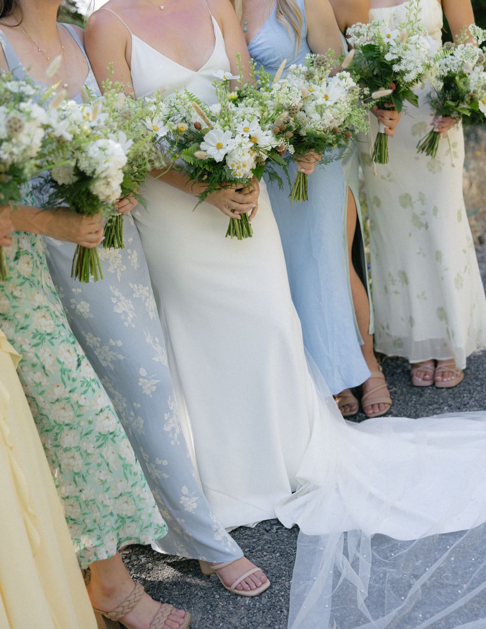 Bridesmaids wearing pastel dresses holding elegant white floral bouquets at an outdoor wedding ceremony.