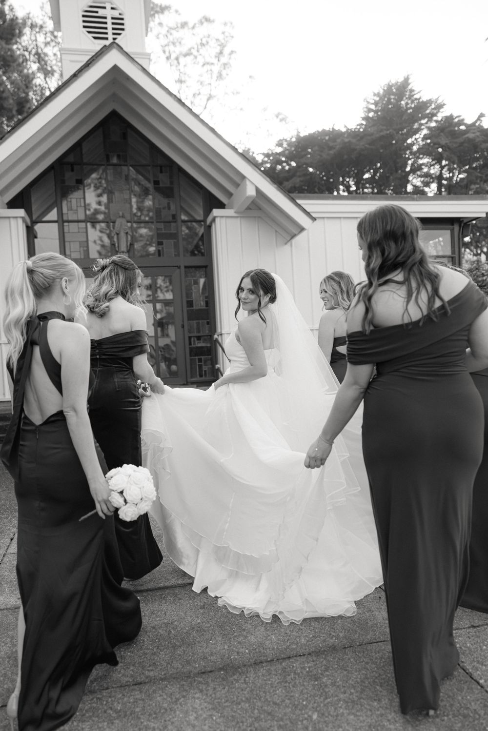 Bridesmaids in black dresses help adjust a flowing white wedding gown outside a small chapel.