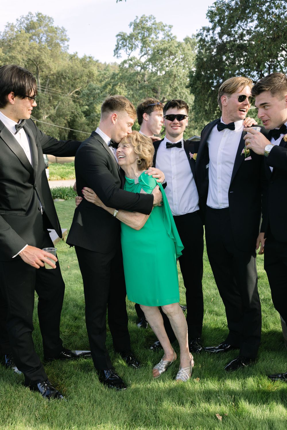 A group in formal black attire and bowties gathered outdoors with one person wearing a bright green dress.