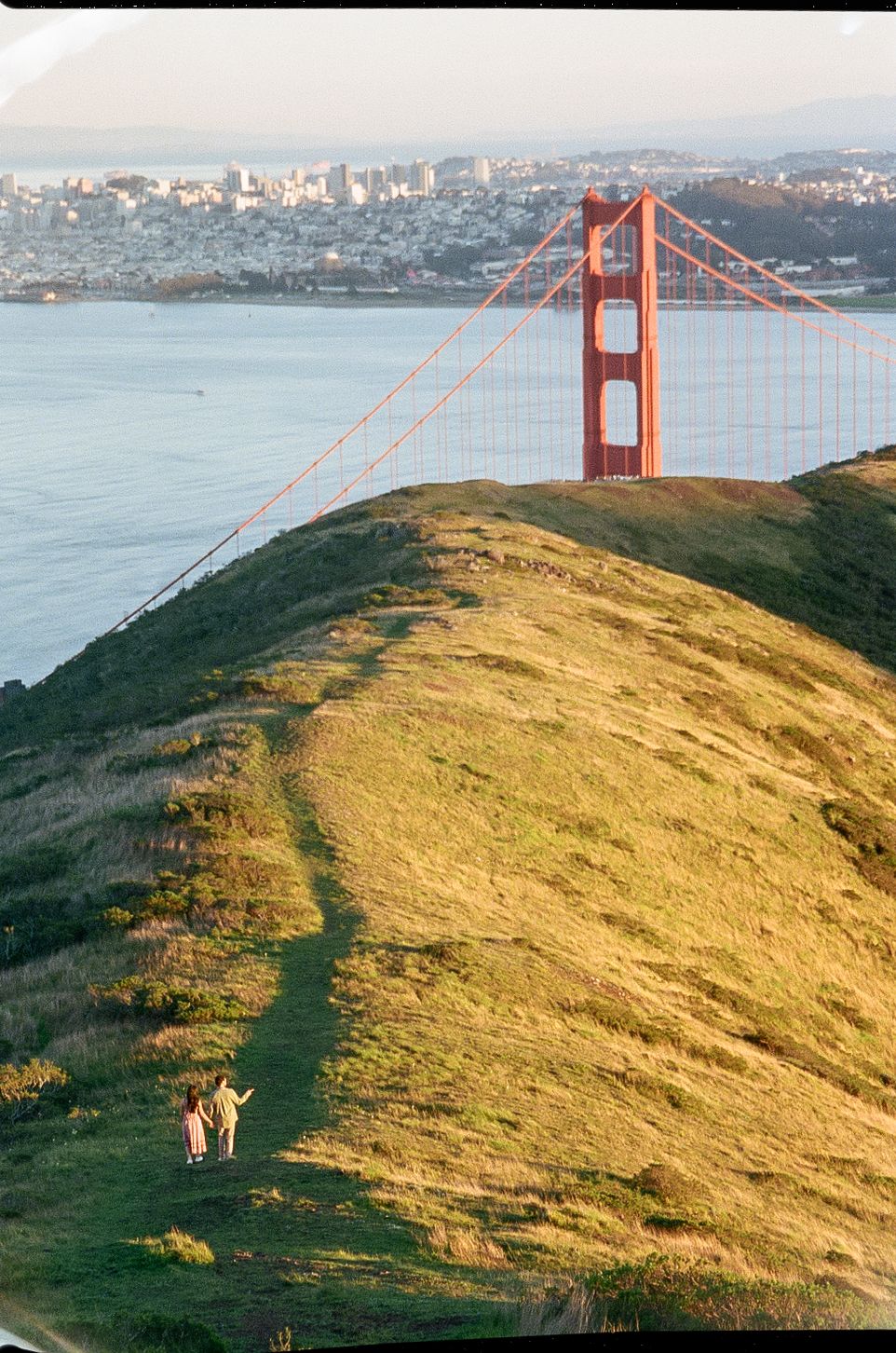 Two people hike on a grassy hillside trail overlooking the iconic Golden Gate Bridge and San Francisco Bay at sunset.