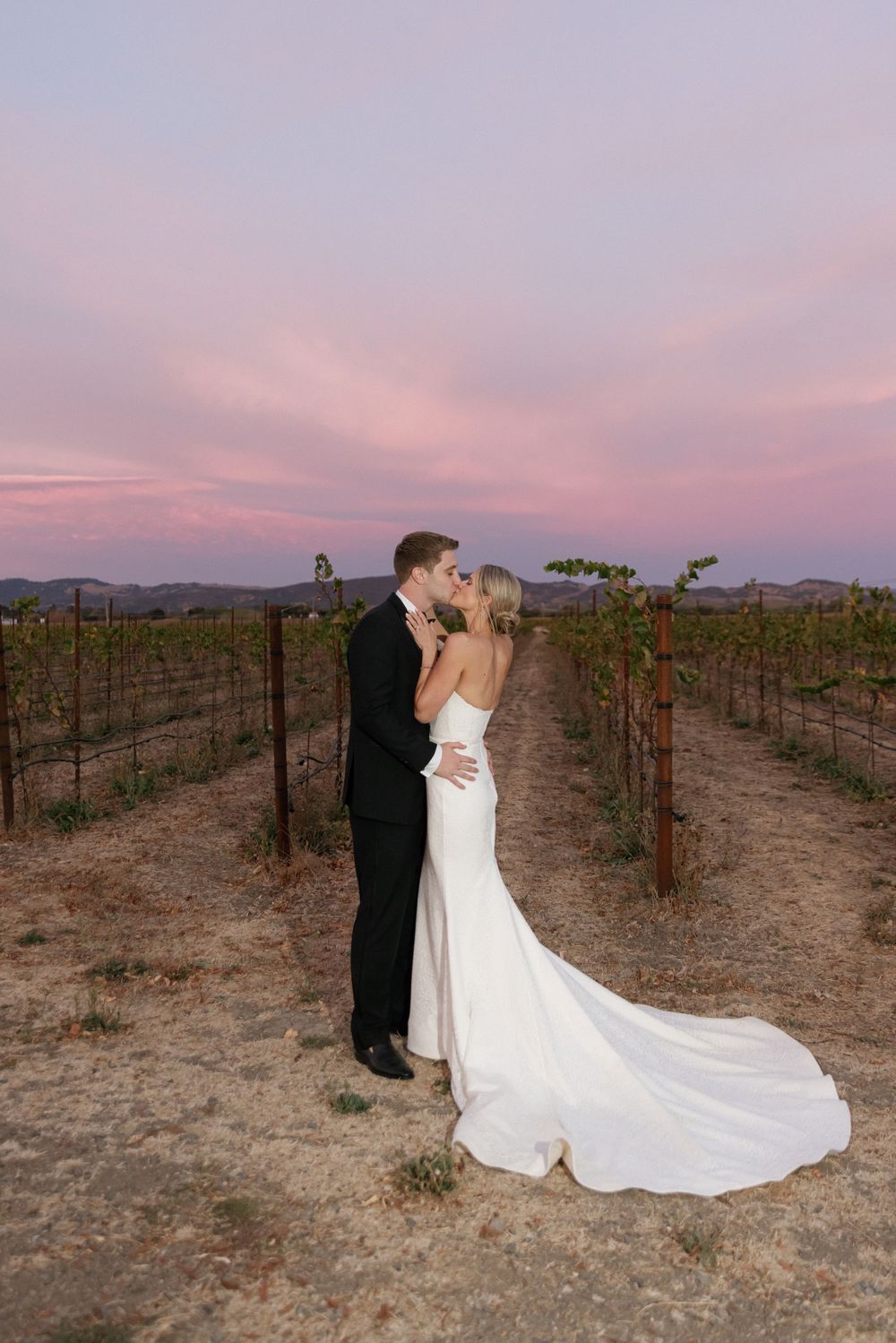 A couple embraces in a vineyard at sunset with mountains in the background and a flowing white wedding dress train.