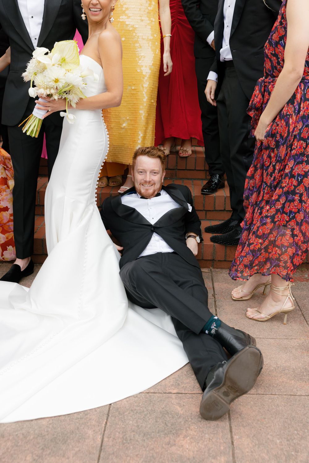 At a formal wedding, a tuxedo-clad guest playfully lies on the ground next to a bride holding a white bouquet.