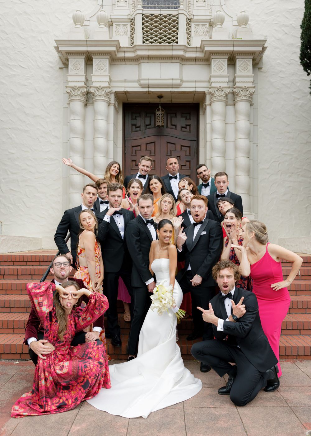 Wedding party poses together on church steps in formal attire with elegant white and red dresses and black tuxedos.