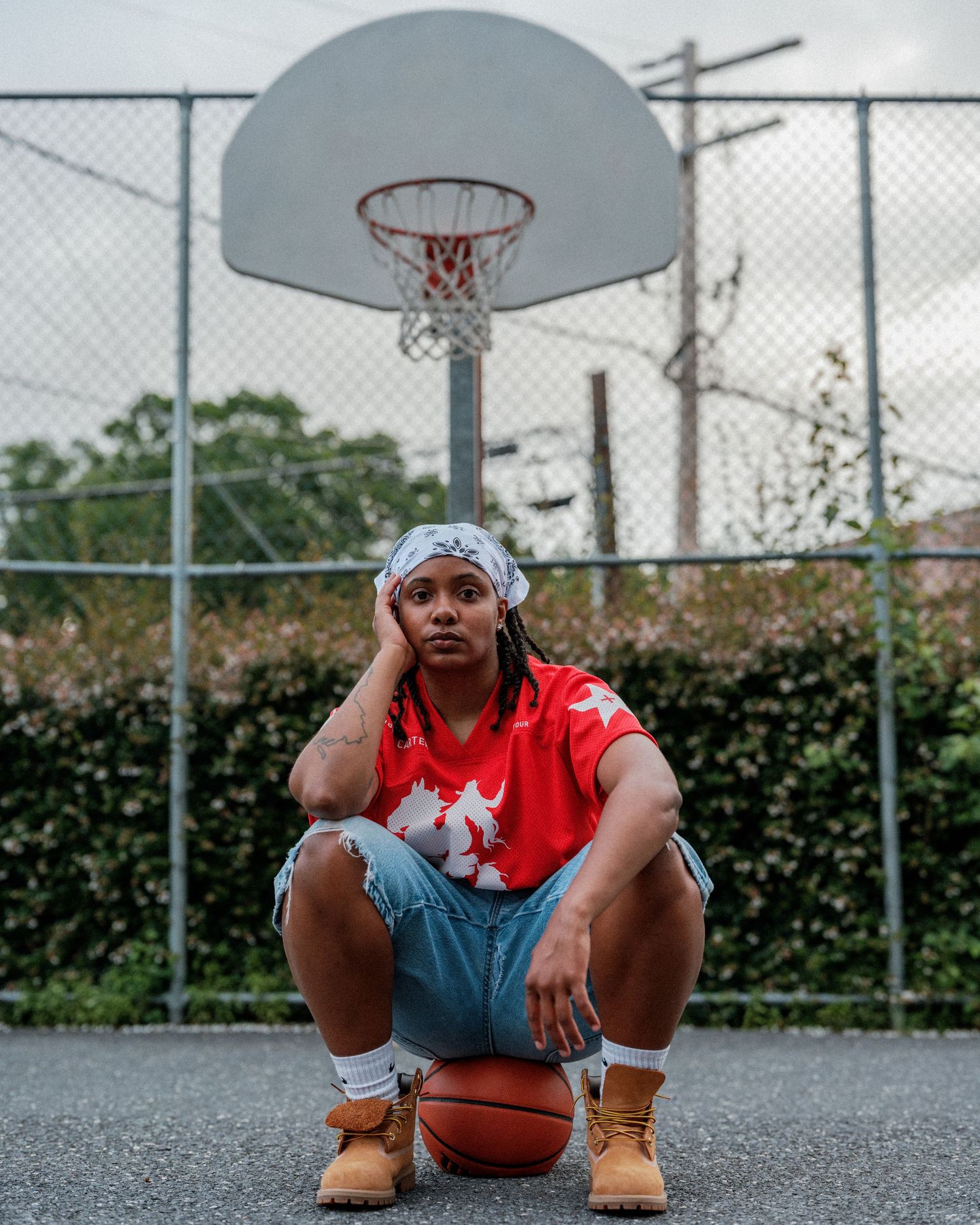 A basketball player in a red jersey and denim shorts sits on a basketball in front of an outdoor court with a hoop.