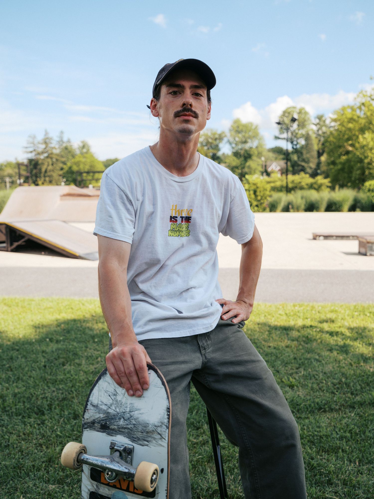 Skateboarder wearing a white t-shirt and cap sits on grass near a skate park on a sunny day.