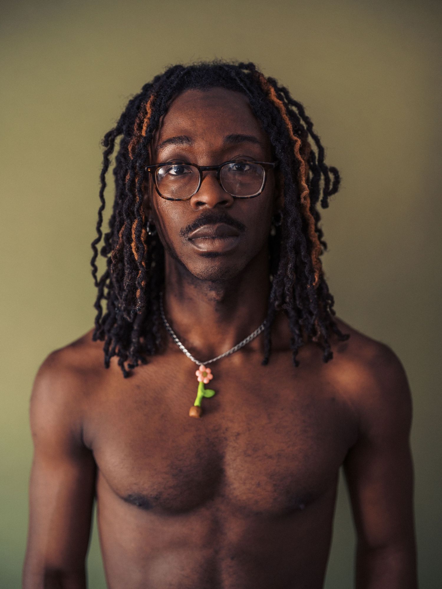 Portrait photograph showing dreadlocked hair, glasses and a necklace with pendant against muted background.
