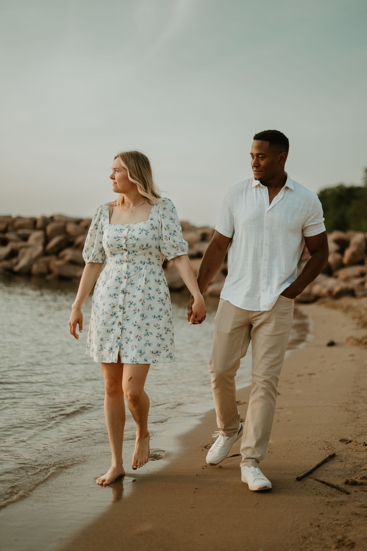 A couple walks hand-in-hand along a sandy beach shore in Milwaukee, woman in floral dress, man in white shirt and khaki pants.