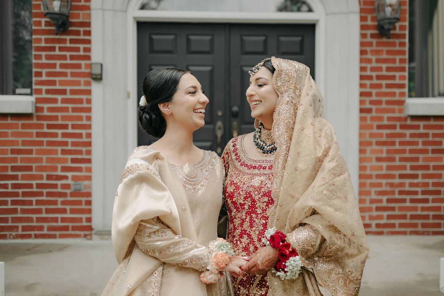 Two laughing friends share a moment outside a brick building while wearing traditional South Asian wedding attire.