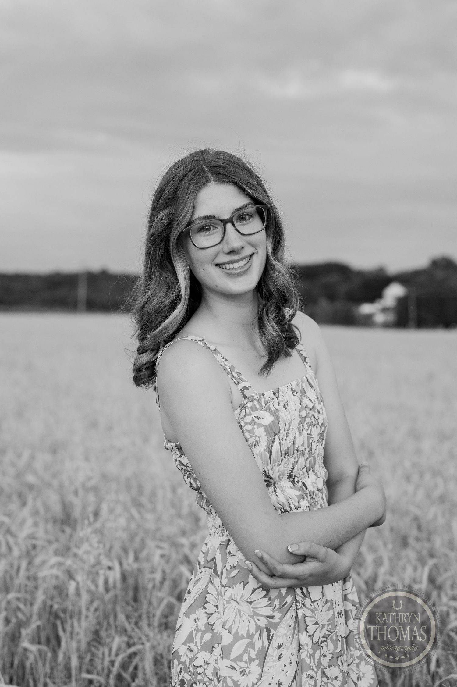 Black and white portrait of someone in floral dress smiling warmly in wheat field.