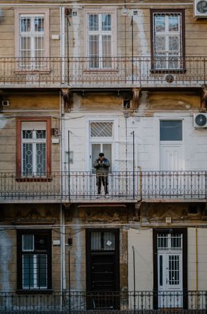 An old European apartment building facade with iron balconies and windows, showing a person standing on a middle floor balcony.