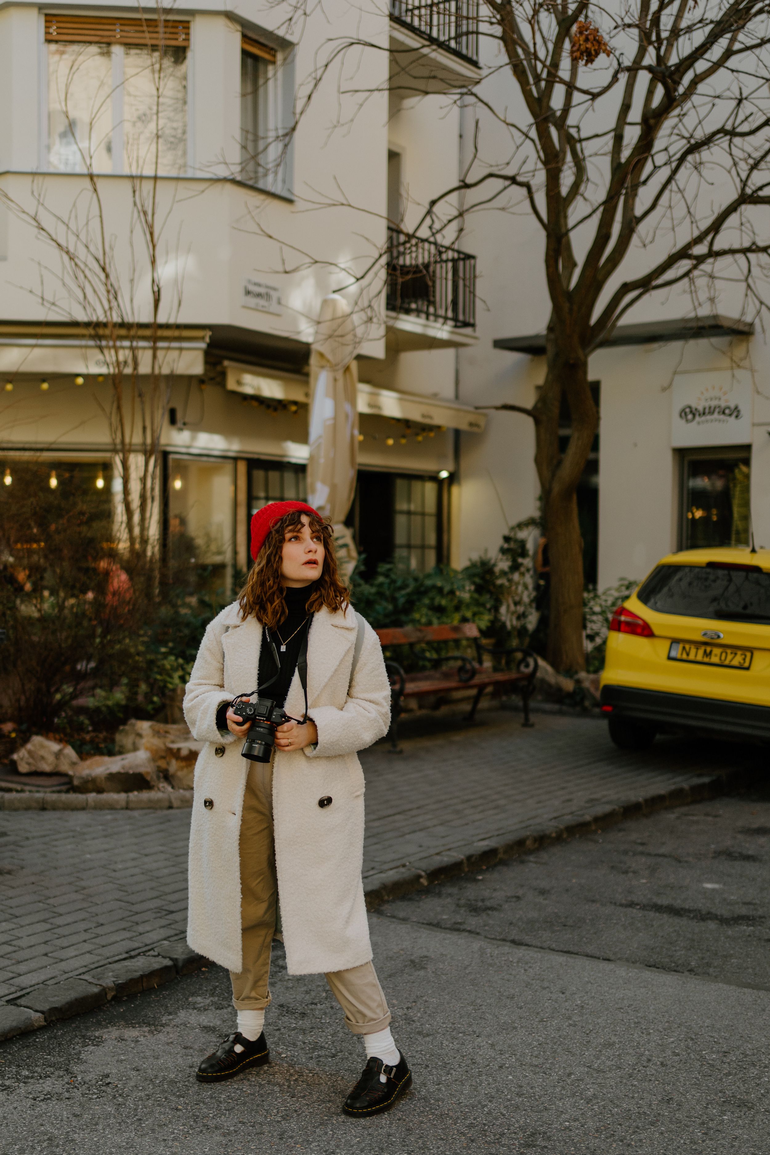 Winter street style with cream coat, red beret and camera on cobblestone street next to yellow taxi in city.