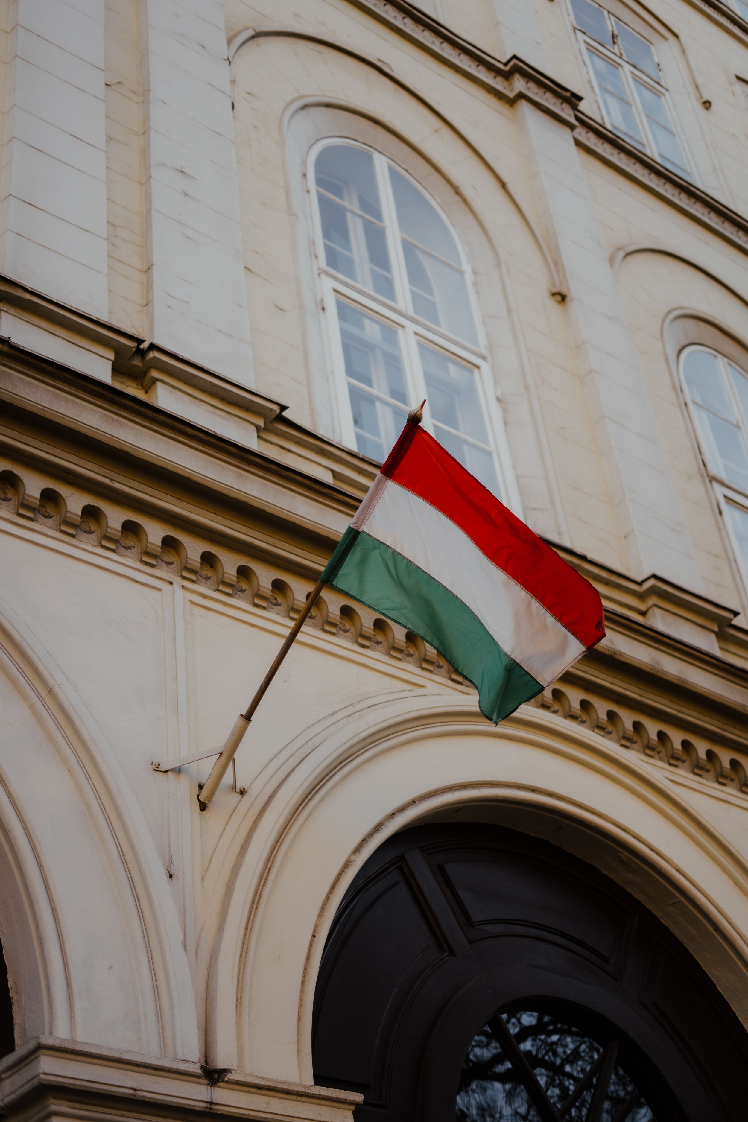 Hungarian flag displayed on classical building facade with arched windows and ornate architectural details.