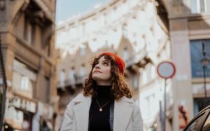 Person wearing a red beret and white coat gazes upward while standing on a European city street in winter.