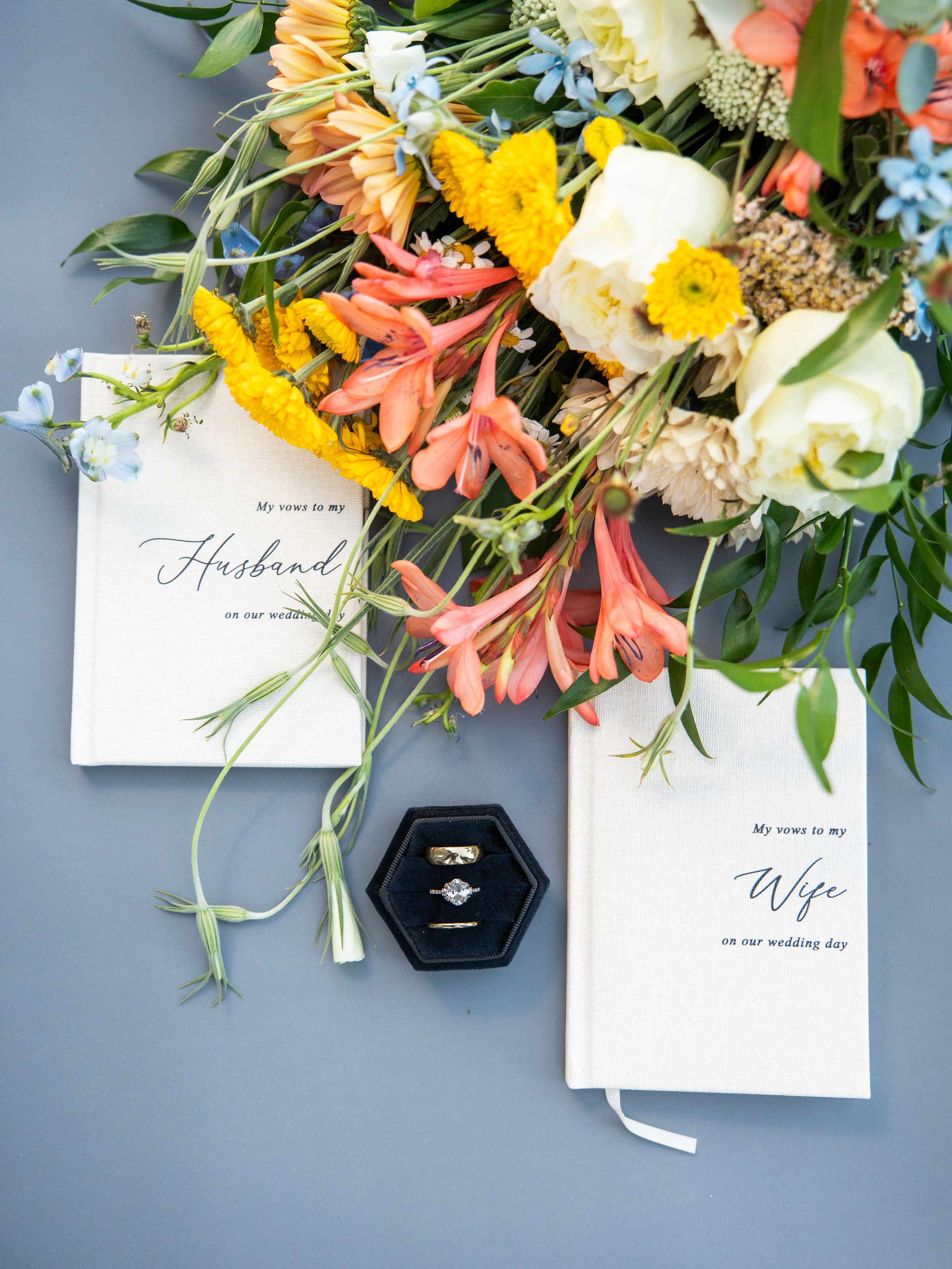 Wedding vows and ring box displayed with colorful spring flowers including yellow daffodils and coral alstroemeria on blue background.
