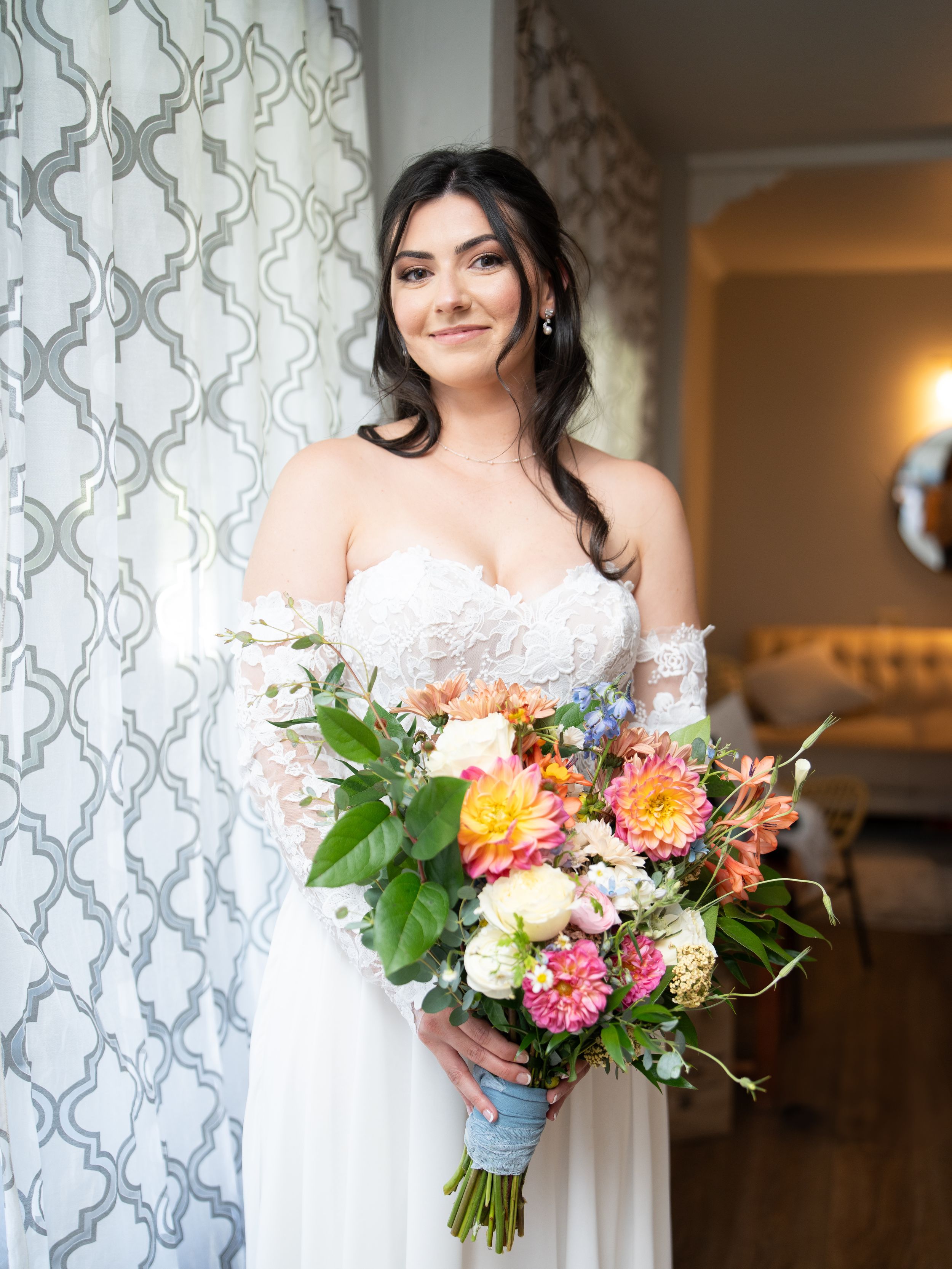 A bridal portrait showing a person in a white lace wedding dress holding a vibrant bouquet of mixed flowers against patterned curtains.