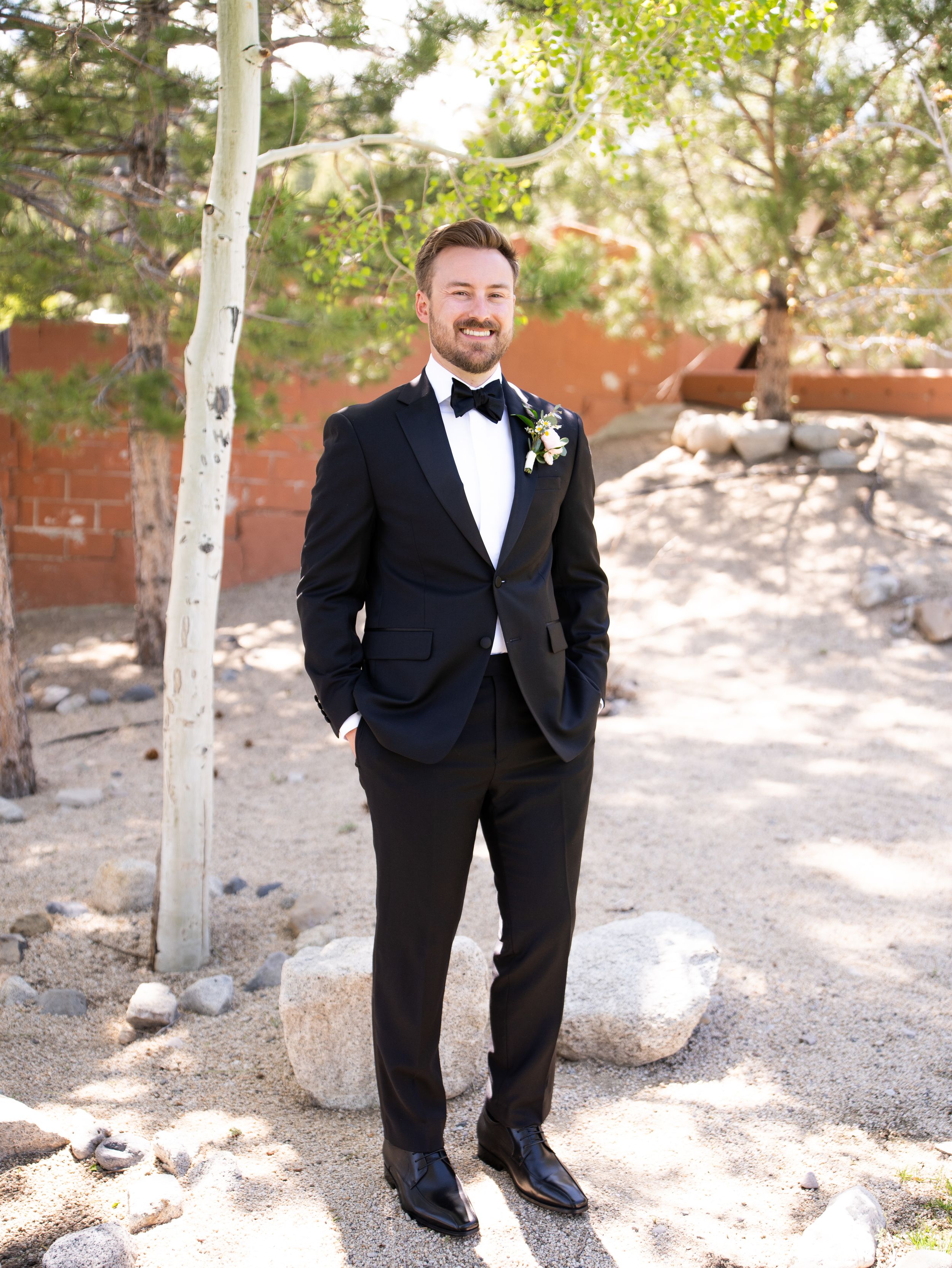 A groom in a classic black tuxedo and bow tie stands outdoors near an aspen tree on his wedding day.