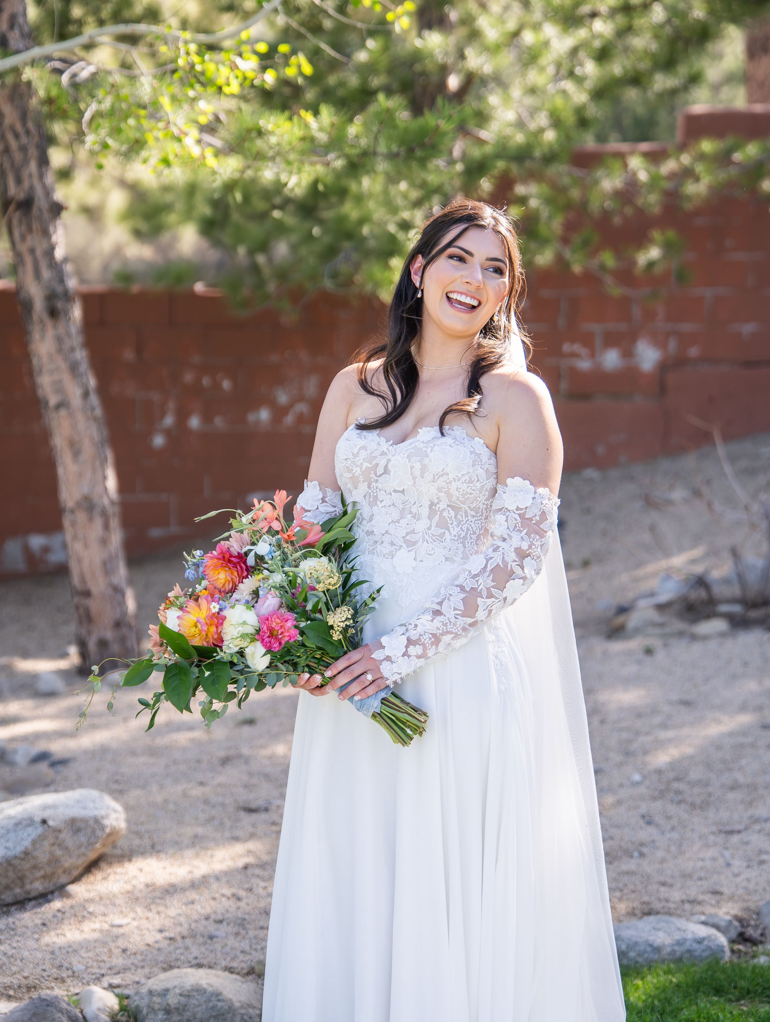 A figure in a flowing white lace wedding gown holds a vibrant bouquet of flowers in an outdoor garden setting.