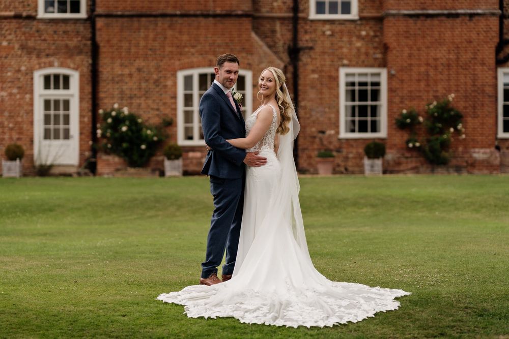 An elegantly dressed married couple stand together on a manicured lawn in front of a historic brick building.