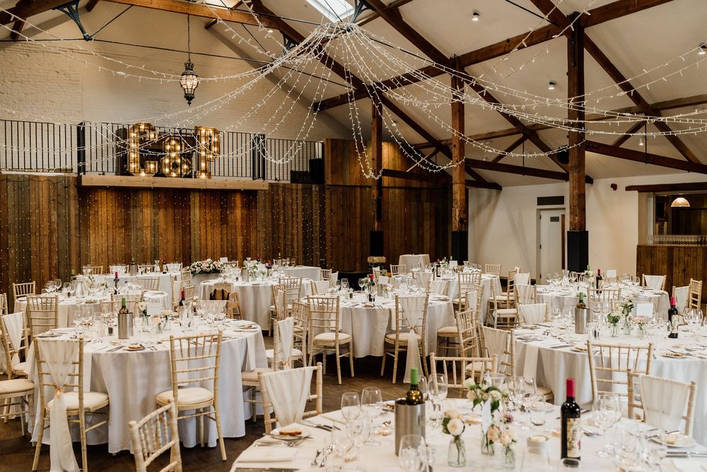 Elegant wedding reception at Oxnead Hall with wooden beams, white tablecloths and chiavari chairs in rustic barn setting.